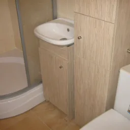 Łazienka z kabiną prysznicową i umywalką z szafką oraz wysoką szafką, wykończone w okleinie imitującej jasne drewno z wyraźnym usłojeniem.